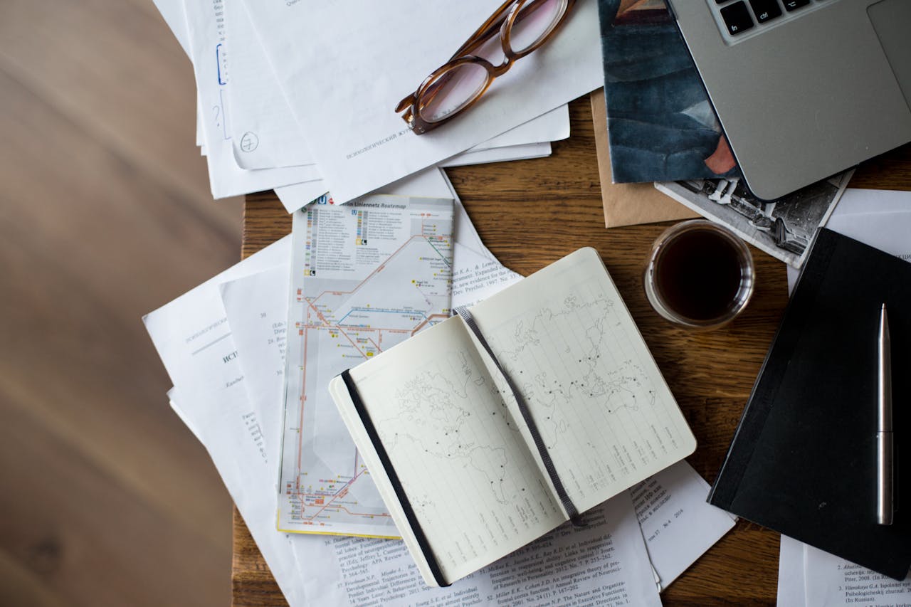 Flat lay of a workspace with coffee, maps, laptop, and documents on a wooden desk.