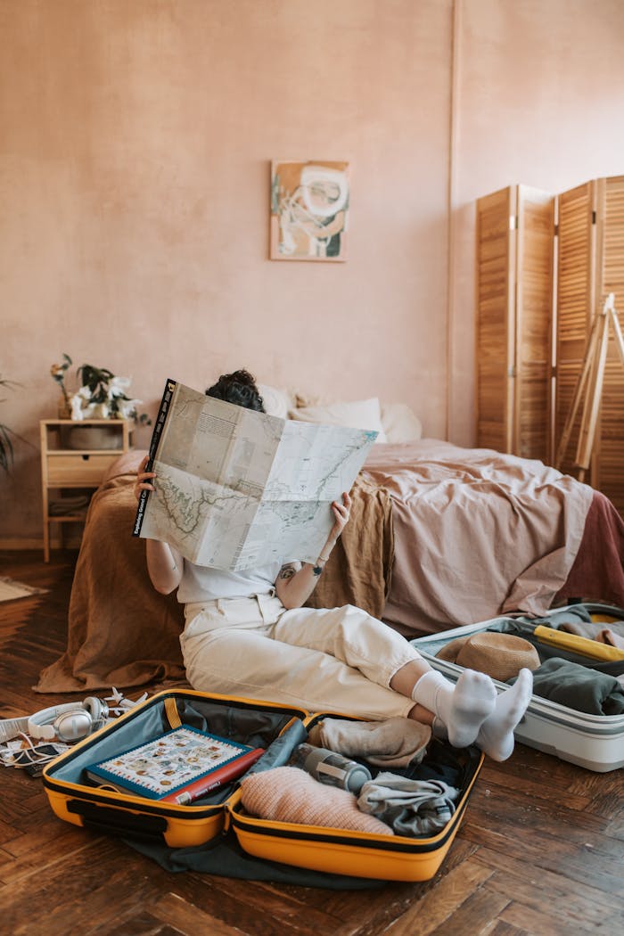 A woman sitting on the floor reading a map, surrounded by travel items and a packed suitcase in a cozy bedroom.